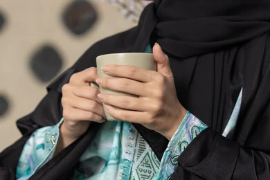 مطاعم  وكافيهات في الامارات العربية المتحدة، قضاء وقت الفراغ باحتساء المشروب المفضل, تناول الأعشاب الشتوية الدافئة، الاستمتاع بالمشروبات الساخنة، صورة مقربة لامراة اماراتية ترتدي العباءة والحجاب تجلس على اريكة مريحة تقوم بشرب القهوة في المقهى