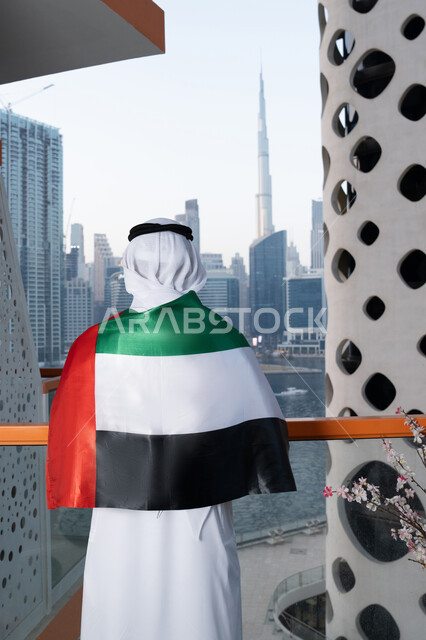 Gestures of pride and admiration, celebrating the anniversary of the independence of the United Arab Emirates, UAE National Day, December 2, a photo from the back of an Emirati Gulf Arab man wearing a kandura and ghutra, placing the UAE flag on his shoulder during the day, walking in the streets