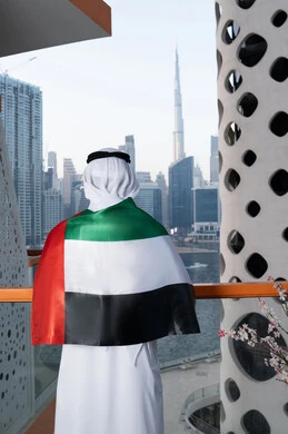 Gestures of pride and admiration, celebrating the anniversary of the independence of the United Arab Emirates, UAE National Day, December 2, a photo from the back of an Emirati Gulf Arab man wearing a kandura and ghutra, placing the UAE flag on his shoulder during the day, walking in the streets