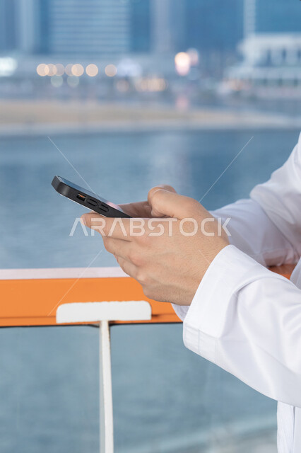 Exchanging conversations, communication and news with family and friends, an Emirati Gulf Arab man wearing a white kandura and ghutra holding a mobile phone, using a modern and advanced technical device, working remotely, browsing applications and social networking programs