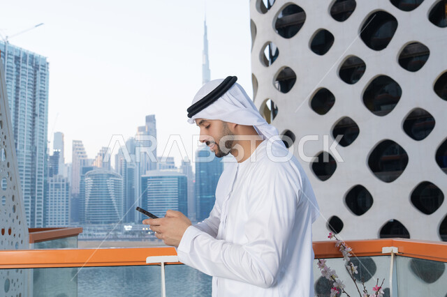 التواصل وتبادل الأحاديث والأخبار مع الأهل والأصدقاء، رجل عربي خليجي اماراتي يرتدي الكندورة والغترة البيضاء يمسك بيده الهاتف المحمول، استخدام جهاز تقني حديث ومتطور، العمل عن بعد، تصفح التطبيقات وبرامج التواصل الاجتماعي
