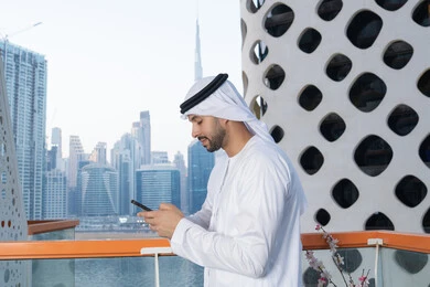 Communicating and exchanging conversations and news with family and friends, an Emirati Gulf Arab man wearing a white kandura and ghutra holding a mobile phone, using a modern and advanced technical device, working remotely, browsing applications and social networking programs