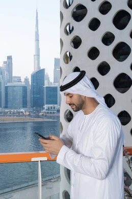 Working remotely, browsing social media applications and programs, communicating and exchanging conversations and news with family and friends, an Emirati Gulf Arab man wearing a white kandura and ghutra holding a mobile phone in his hand, using a modern and advanced technical device