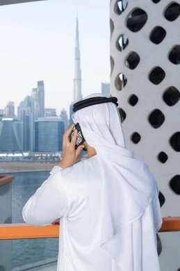 Using a modern and advanced technical device, an Arab Gulf Emirati man wearing a white kandura and ghutra holding a mobile phone, completing tasks and working remotely, communicating and exchanging conversations with family and friends, making a phone call