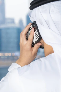انجاز المهام والعمل عن بعد، التواصل وتبادل الأحاديث مع الأهل والأصدقاء، اجراء مكالمة هاتفية، استخدام جهاز تقني حديث ومتطور،  رجل عربي خليجي اماراتي يرتدي الكندورة والغترة البيضاء يمسك بيده الهاتف المحمول
