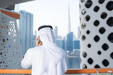 Accomplishing tasks and working remotely, communicating and exchanging conversations with family and friends, making a phone call, using a modern and advanced technical device, an Arab Gulf Emirati man wearing a kandura and a white ghutra holding a mobile phone in his hand