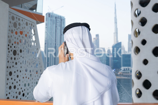 Using a modern and advanced technical device, an Arab Gulf Emirati man wearing a white kandura and ghutra holding a mobile phone, completing tasks and working remotely, communicating and exchanging conversations with family and friends, making a phone call