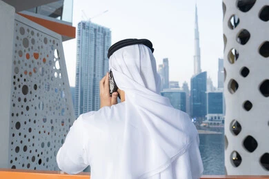 Using a modern and advanced technical device, an Arab Gulf Emirati man wearing a white kandura and ghutra holding a mobile phone, completing tasks and working remotely, communicating and exchanging conversations with family and friends, making a phone call