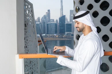 Accomplishing work tasks remotely, managing and organizing business affairs via laptop, gestures of happiness and pleasure, using a modern and advanced technical device, an Arab Gulf Emirati man wearing a white kandura and ghutra using a laptop