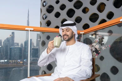 قضاء وقت الفراغ باحتساء المشروب المفضل، تناول الأعشاب الشتوية الدافئة، الاستمتاع بالمشروبات الساخنة، رجل عربي خليجي اماراتي يرتدي الكندورة والغترة البيضاء  يقوم بشرب القهوة في المقهى، إيماءات السعادة والسرور