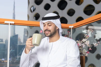 الاستمتاع بالمشروبات الساخنة، رجل عربي خليجي اماراتي يرتدي الكندورة والغترة البيضاء  يقوم بشرب القهوة في المقهى، إيماءات السعادة والسرور، قضاء وقت الفراغ باحتساء المشروب المفضل، تناول الأعشاب الشتوية الدافئة