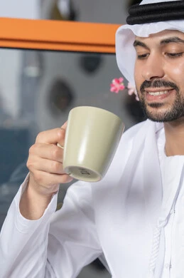 قضاء وقت الفراغ باحتساء المشروب المفضل، تناول الأعشاب الشتوية الدافئة، الاستمتاع بالمشروبات الساخنة، رجل عربي خليجي اماراتي يرتدي الكندورة والغترة البيضاء  يقوم بشرب القهوة في المقهى، إيماءات السعادة والسرور