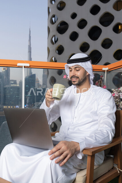 الاستمتاع بالمشروب المفضل، إنجاز مهام العمل عن بعد، إدارة شؤون الأعمال وتنظيمها عبر اللابتوب، ايماءات السعادة والسرور، استخدام جهاز تقني حديث ومتطور،رجل عربي خليجي اماراتي يرتدي الكندورة والغترة البيضاء يستخدم الحاسوب المحمول ويحمل بيده كوب قهوة