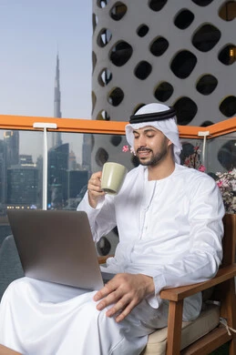 الاستمتاع بالمشروب المفضل، إنجاز مهام العمل عن بعد، إدارة شؤون الأعمال وتنظيمها عبر اللابتوب، ايماءات السعادة والسرور، استخدام جهاز تقني حديث ومتطور،رجل عربي خليجي اماراتي يرتدي الكندورة والغترة البيضاء يستخدم الحاسوب المحمول ويحمل بيده كوب قهوة