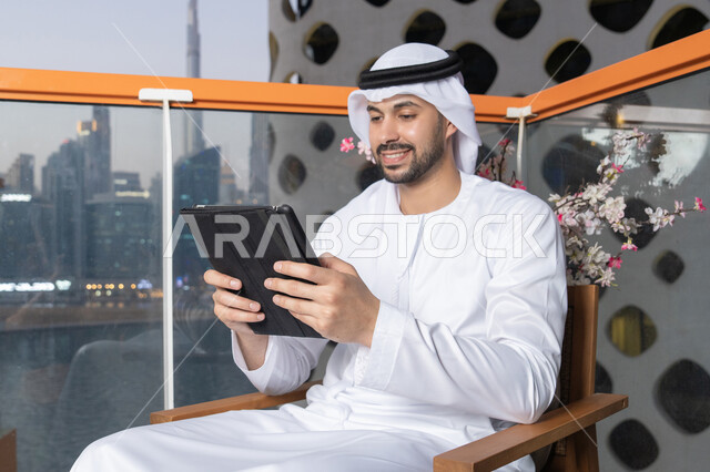 ايماءات السرور والسعادة، استخدام جهاز تقني حديث ومتطور، رجل عربي خليجي اماراتي يرتدي الكندورة والغترة البيضاء يستخدم الجهاز اللوحي، إنجاز مهام العمل عن بعد، إدارة شؤون الأعمال وتنظيمها عبر الايباد
