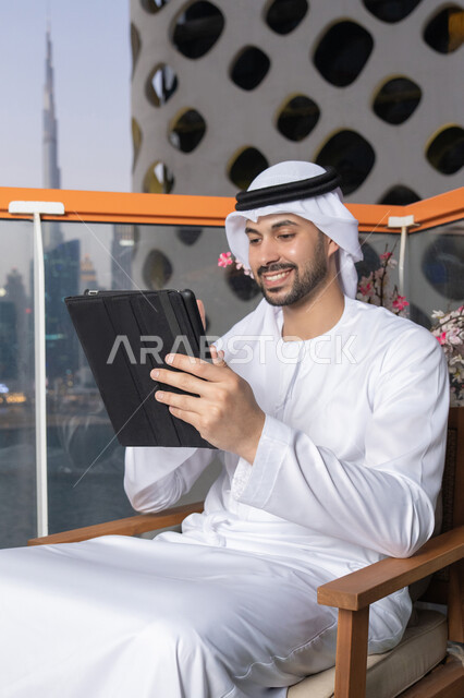رفع كف اليد بايماءات القاء التحية والسلام، إجراء مكالمة فيديو، إنجاز مهام العمل عن بعد، إدارة شؤون الأعمال وتنظيمها عبر الايباد، استخدام جهاز تقني حديث ومتطور، رجل عربي خليجي اماراتي يرتدي الكندورة والغترة البيضاء يستخدم الجهاز اللوحي