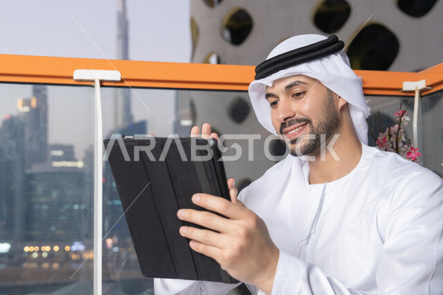 إجراء مكالمة فيديو، إنجاز مهام العمل عن بعد، إدارة شؤون الأعمال وتنظيمها عبر الايباد، استخدام جهاز تقني حديث ومتطور، رجل عربي خليجي اماراتي يرتدي الكندورة والغترة البيضاء يستخدم الجهاز اللوحي، رفع كف اليد بايماءات القاء التحية والسلام
