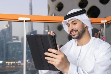 إجراء مكالمة فيديو، إنجاز مهام العمل عن بعد، إدارة شؤون الأعمال وتنظيمها عبر الايباد، استخدام جهاز تقني حديث ومتطور، رجل عربي خليجي اماراتي يرتدي الكندورة والغترة البيضاء يستخدم الجهاز اللوحي، رفع كف اليد بايماءات القاء التحية والسلام