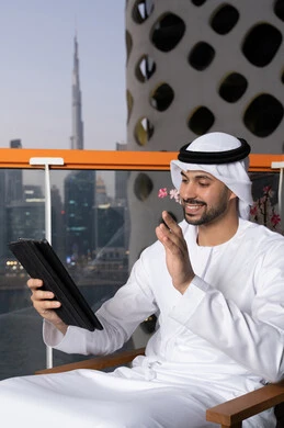 Raising the palm of the hand in greeting gestures, making a video call, completing work tasks remotely, managing and organizing business affairs via iPad, using a modern and advanced technical device, a close-up of an Emirati Gulf Arab man wearing a white kandura and ghutra using a tablet