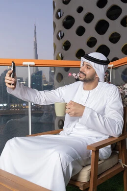 توثيق اللحظات السعيدة، التقاط الصور الشخصية باستخدام الجوال، استخدام جهاز تقني حديث ومتطور في تخليد الذكريات، رجل عربي خليجي اماراتي يرتدي الكندورة والغترة البيضاء يمسك الهاتف المحمول ويأخذ صورة سيلفي بإيماءات السرور، احتساء مشروب القهوة