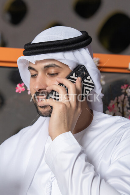استخدام جهاز تقني حديث ومتطور،  رجل عربي خليجي اماراتي يرتدي الكندورة والغترة البيضاء يمسك بيده الهاتف المحمول، انجاز المهام والعمل عن بعد، التواصل وتبادل الأحاديث مع الأهل والأصدقاء، اجراء مكالمة هاتفية