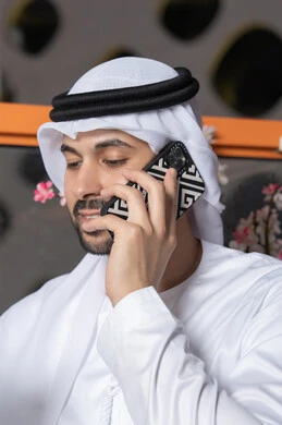 استخدام جهاز تقني حديث ومتطور،  رجل عربي خليجي اماراتي يرتدي الكندورة والغترة البيضاء يمسك بيده الهاتف المحمول، انجاز المهام والعمل عن بعد، التواصل وتبادل الأحاديث مع الأهل والأصدقاء، اجراء مكالمة هاتفية