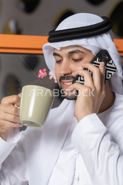 Making a phone call, using a modern and advanced technical device, drinking your favorite drink, an Emirati Gulf Arab man wearing a white kandura and ghutra holding a mobile phone and a cup of coffee, working remotely, communicating and exchanging conversations with family and friends