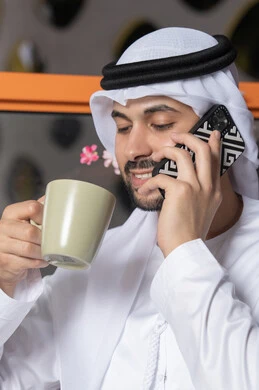 اجراء مكالمة هاتفية، استخدام جهاز تقني حديث ومتطور، احتساء المشروب المفضل، رجل عربي خليجي اماراتي يرتدي الكندورة والغترة البيضاء يمسك بيده الهاتف المحمول وكوب من القهوة، العمل عن بعد،  التواصل وتبادل الأحاديث مع الأهل والأصدقاء