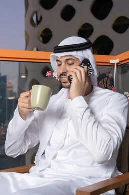 Making a phone call, using a modern and advanced technical device, drinking your favorite drink, an Emirati Gulf Arab man wearing a white kandura and ghutra holding a mobile phone and a cup of coffee, working remotely, communicating and exchanging conversations with family and friends