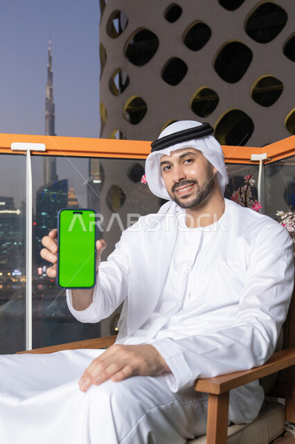 Empty green chroma key screen display, integrating technology into work life, an Emirati Gulf Arab man wearing a white kandura and ghutra holding a mobile phone, using modern technology to accomplish work tasks