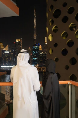 Looking at the towers and skyscrapers, the growth and urban development of facilities in the Emirates, the concept of love and intimacy between spouses, interest in outward appearance, an Arab Gulf Emirati man wearing a white kandura and ghutra standing with his wife