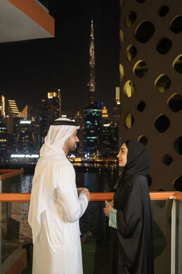 Looking at the towers and skyscrapers, the growth and urban development of facilities in the Emirates, the concept of love and intimacy between spouses, interest in outward appearance, an Arab Gulf Emirati man wearing a white kandura and ghutra standing with his wife