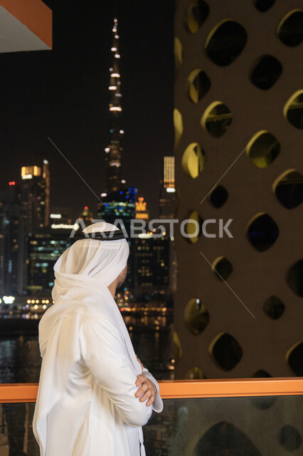 The concept of concern for outward appearance, masculinity and elegance, an Arab Gulf Emirati man wearing a white kandura and ghutra standing straight with his hands crossed, looking at the towers and skyscrapers, the growth and urban development of facilities in the Emirates
