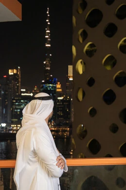 مفهوم الاهتمام بالمظهر الخارجي و الرجولة والأناقه، رجل عربي خليجي إماراتي يرتدي الكندورة والغترة البيضاء يقف باستقامة بايدي متقاطعه, النظر الى الابراج وناطحات السحاب، النمو والتقدم العمراني للمنشآت في الامارات