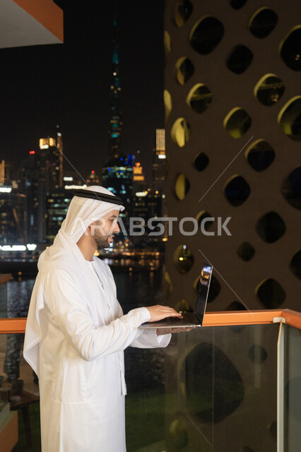 إنجاز مهام العمل عن بعد، إدارة شؤون الأعمال وتنظيمها عبر اللابتوب، ايماءات السعادة والسرور، استخدام جهاز تقني حديث ومتطور، رجل عربي خليجي اماراتي يرتدي الكندورة والغترة البيضاء يستخدم الحاسوب المحمول