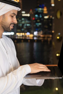 إدارة شؤون الأعمال وتنظيمها عبر اللابتوب، ايماءات السعادة والسرور، استخدام جهاز تقني حديث ومتطور، رجل عربي خليجي اماراتي يرتدي الكندورة والغترة البيضاء يستخدم الحاسوب المحمول، إنجاز مهام العمل عن بعد