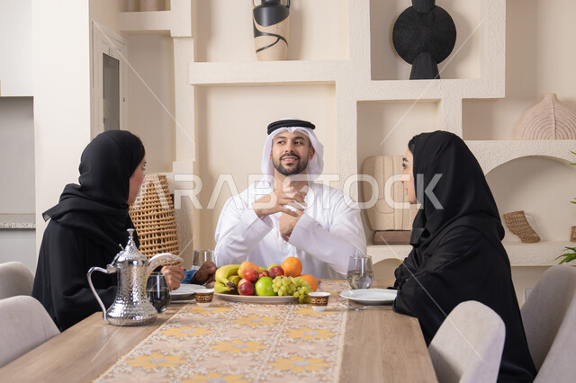 تبادل الأخبار والأحاديث، حسن إكرام الضيف، قضاء وقت عائلي ممتع، سناك خفيف ومنعش، موسم الفاكهة الصحية اللذيذة، التفاهم والمحبة بين الزوجين، اجواء الأسرة السعيدة، عائلة عربية خليجية اماراتية يجلسون خلف الطاولة في المنزل، تناول الفواكه الطازجة بإيماءات السعادة