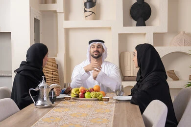 تبادل الأخبار والأحاديث، حسن إكرام الضيف، قضاء وقت عائلي ممتع، سناك خفيف ومنعش، موسم الفاكهة الصحية اللذيذة، التفاهم والمحبة بين الزوجين، اجواء الأسرة السعيدة، عائلة عربية خليجية اماراتية يجلسون خلف الطاولة في المنزل، تناول الفواكه الطازجة بإيماءات السعادة