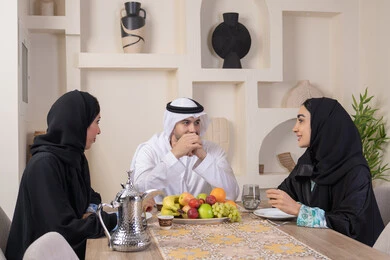 مفهوم حسن إكرام الضيف، قضاء وقت عائلي ممتع، سناك خفيف ومنعش، موسم الفاكهة الصحية اللذيذة، التفاهم والمحبة بين الزوجين، اجواء الأسرة السعيدة، عائلة عربية خليجية اماراتية يجلسون خلف الطاولة في المنزل، تناول الفواكه الطازجة بإيماءات السعادة والسرور