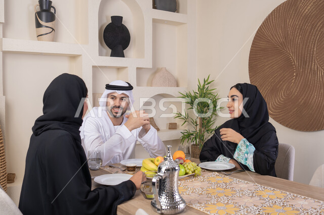 شرب القهوة العربية، تبادل الأخبار والأحاديث، حسن إكرام الضيف، قضاء وقت عائلي ممتع، موسم الفاكهة الصحية اللذيذة، التفاهم والمحبة بين الزوجين، اجواء الأسرة السعيدة، عائلة عربية خليجية اماراتية يجلسون خلف الطاولة في المنزل، تناول الفواكه الطازجة بإيماءات السعادة