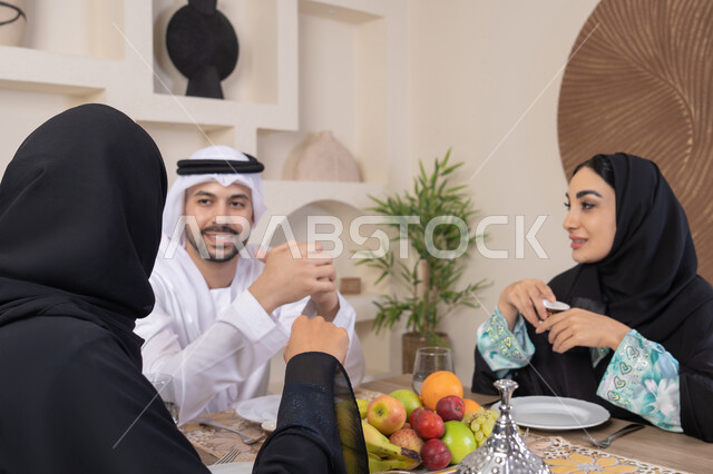 Drinking Arabic coffee, exchanging news and conversations, treating guests well, spending quality family time, the season of delicious healthy fruits, understanding and love between spouses, a happy family atmosphere, an Arab Gulf Emirati family sitting behind the table at home, eating fresh fruits with gestures of happiness