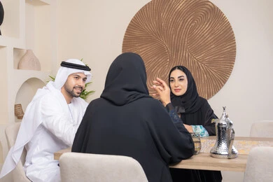 تعابير السعادة والسرور، جلسة وناسة بين الأصدقاء، شرب القهوة العربية، تبادل الأخبار والأحاديث، حسن إكرام الضيف، قضاء وقت عائلي ممتع، اجواء الأسرة السعيدة، عائلة عربية خليجية اماراتية يجلسون خلف الطاولة في المنزل، تناول القهوة العربية الساخنة