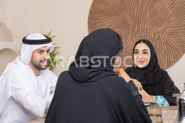 Expressions of happiness and pleasure, a pleasant session among friends, drinking Arabic coffee, exchanging news and conversations, treating guests well, spending enjoyable family time, a happy family atmosphere, an Arab Gulf Emirati family sitting behind a table at home, drinking hot Arabic coffee