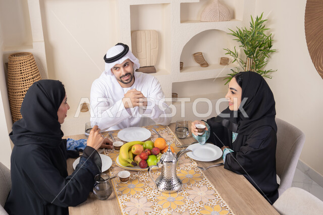 Drinking Arabic coffee, exchanging news and conversations, treating guests well, spending quality family time, the season of delicious healthy fruits, understanding and love between spouses, a happy family atmosphere, an Arab Gulf Emirati family sitting behind the table at home, eating fresh fruits with gestures of happiness