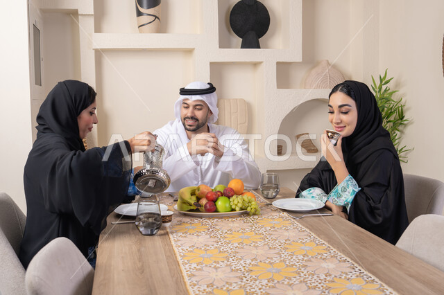 العادات والتقاليد الأصيلة في تقديم القهوة، تناول الفواكه الصحية الطازجة، زوجان عربيان خليجيان اماراتيان يجلسان خلف الطاولة في البيت بإيماءات السعادة والسرور، مفهوم المحبة والتفام بين الزوجين، سيدة محجبة ترتدي العباءة تقوم بصب فنجان من القهوة