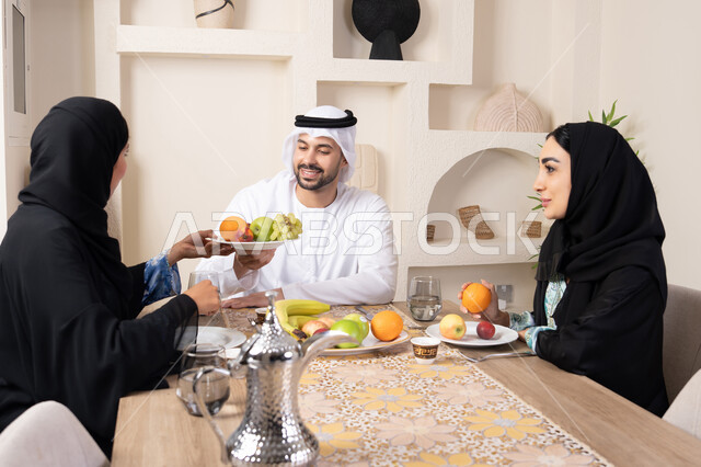 Good hospitality, spending enjoyable family time, a light and refreshing snack, the season of delicious healthy fruits, understanding and love between spouses, a happy family atmosphere, an Arab Gulf Emirati family sitting behind the table at home eating fresh fruits with gestures of happiness and pleasure