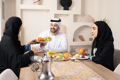حسن إكرام الضيف، قضاء وقت عائلي ممتع، سناك خفيف ومنعش، موسم الفاكهة الصحية اللذيذة، التفاهم والمحبة بين الزوجين، اجواء الأسرة السعيدة، عائلةة عربية خليجية اماراتية يجلسون خلف الطاولة في المنزل يقومان بتناول الفواكه الطازجة بإيماءات السعادة والسرور