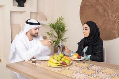 تناول الفواكه الصحية الطازجة، زوجان عربيان خليجيان اماراتيان يجلسان خلف الطاولة في البيت يقومان بشرب القهوة العربية الساخنة بإيماءات السعادة والسرور، مفهوة المحبة والتفام بين الزوجين، جلسة هادئة في المنزل، سيدة محجبة ترتدي العباءة تقوم بصب فنجان من القهوة