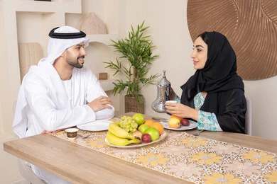 A quiet session at home, eating fresh healthy fruits, an Arab Gulf Emirati couple sitting behind the table at home drinking hot Arabic coffee with gestures of happiness and pleasure, the love and understanding between the spouses is understood, a veiled woman wearing an abaya pouring a cup of coffee
