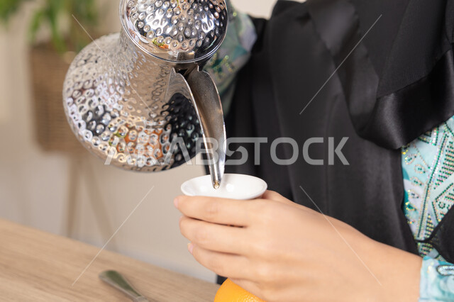 العادات والتقاليد الأصيلة في تقديم القهوة العربية، تصوير مقرب ليد امرأة عربية خليجية اماراتية محجبة ترتدي عباءة سوداء قوم بصب القهوة العربية الساخنة من الدلة الفضية، الاستمتاع لابشرب المشروبات الساخنة